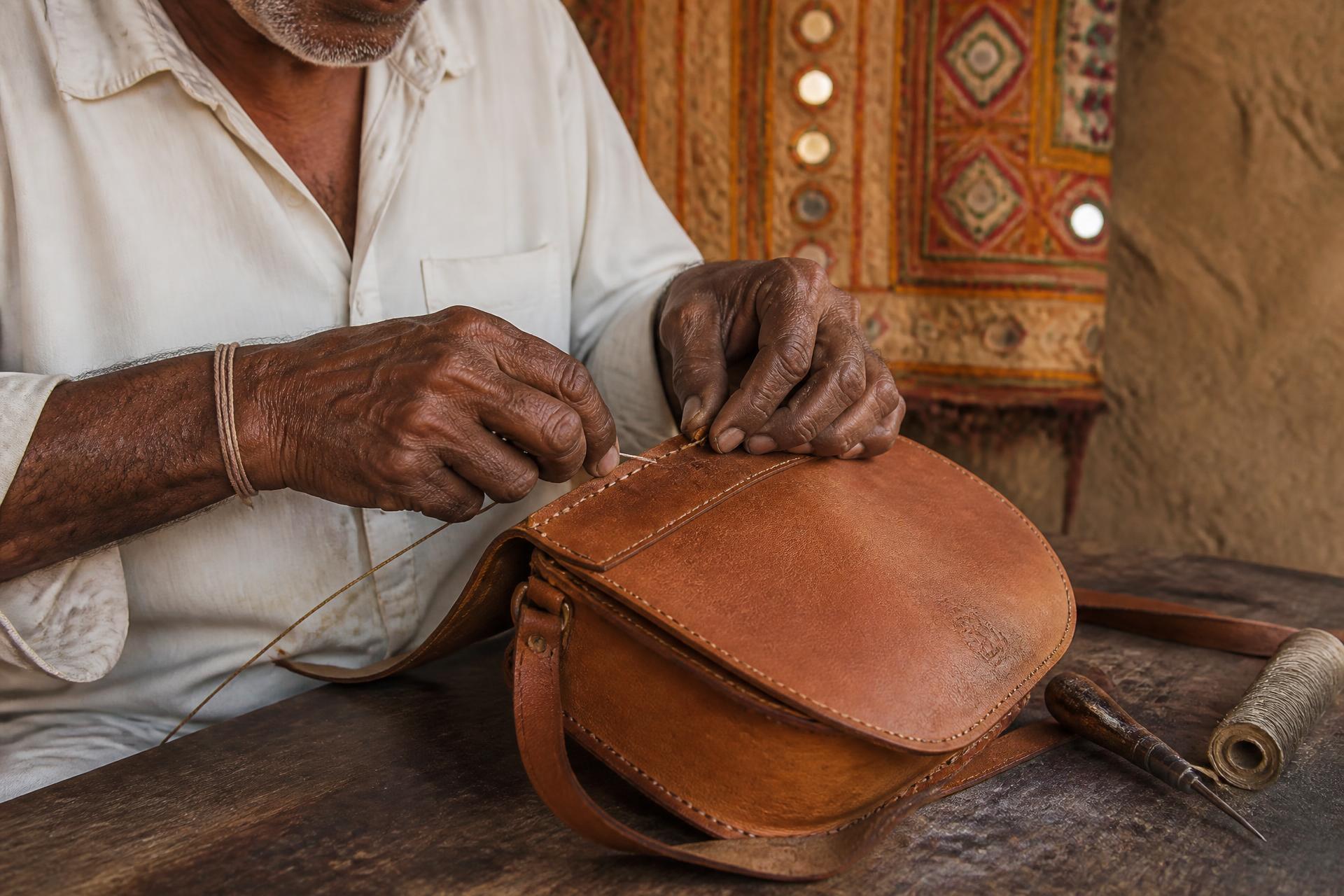 An artisan in Kutch hand-stitching a tan leather sling bag on a wooden workbench